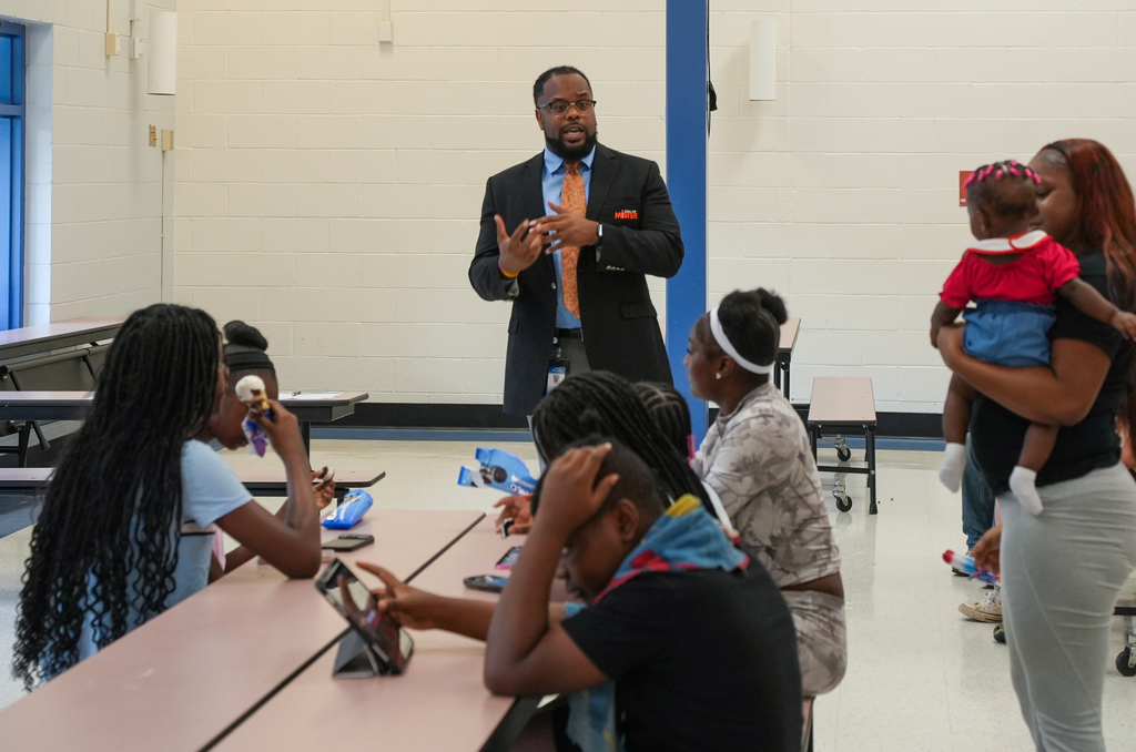 A man in a suit stands in front of a seated group of women and children.