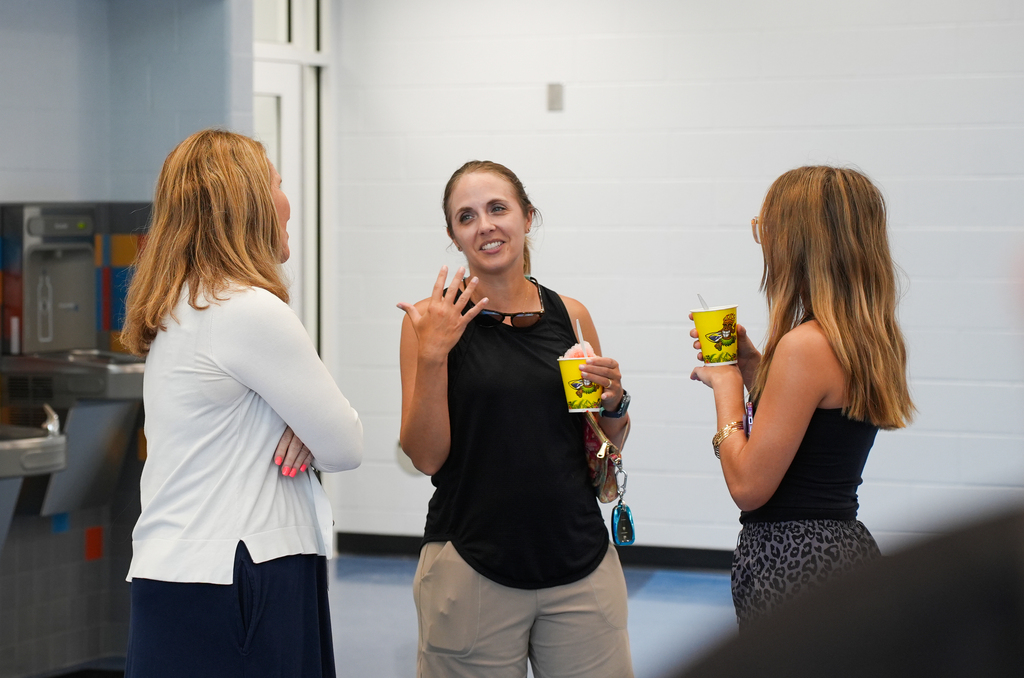 Three women stand in a room, two holding cups, one gesturing with her hand. The woman on the left wears a white shirt, and the others wear black tops.