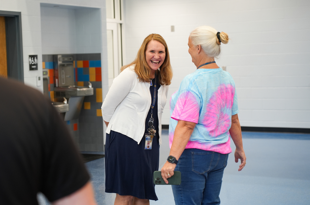Two women converse in a hallway, one wearing a navy skirt and white cardigan, the other in a tie-dye shirt and jeans.
