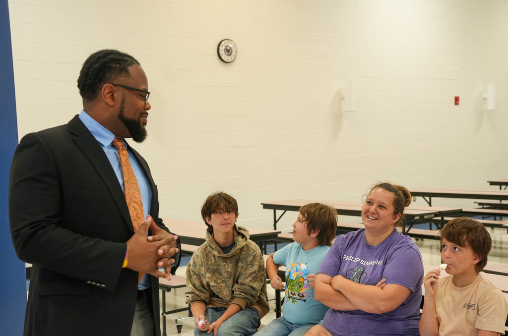 A man in a suit speaks to a group of people sitting at a table. The group includes children and an adult woman.