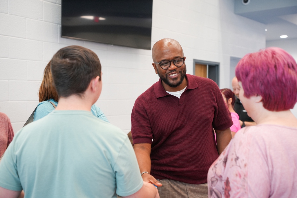 A man wearing glasses stands in a group, conversing with a woman with pink hair and another person.