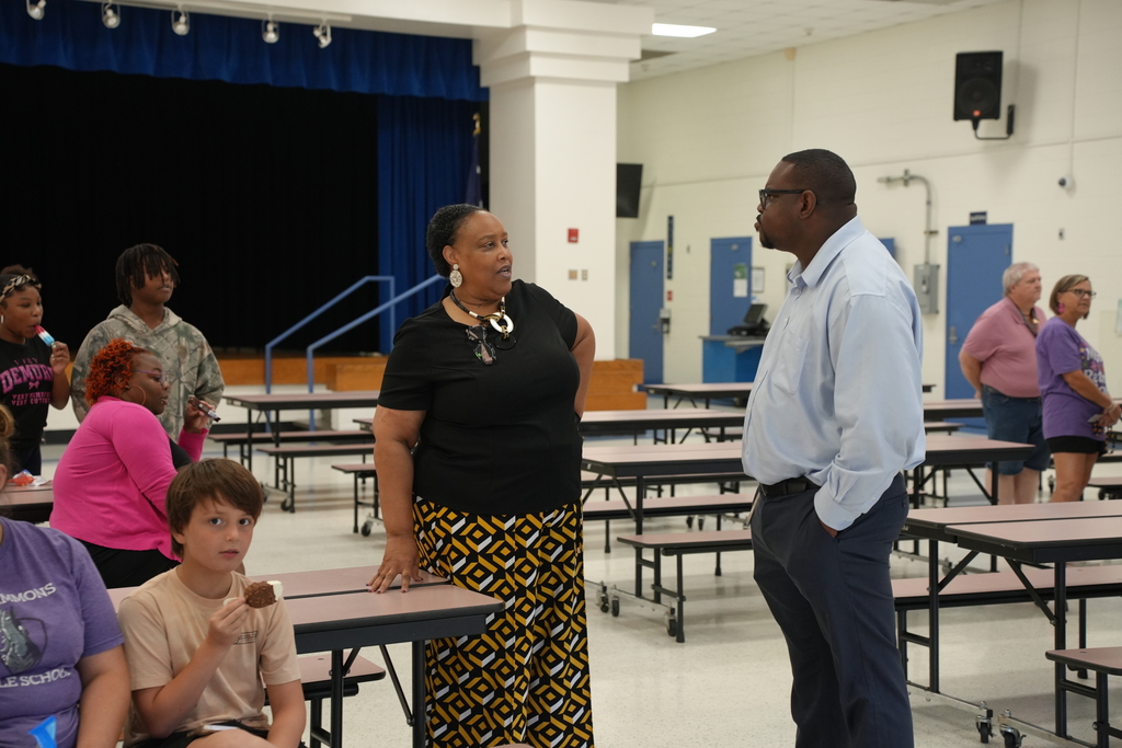 In a room, a man and woman converse near a table. Several people sit or stand around tables.