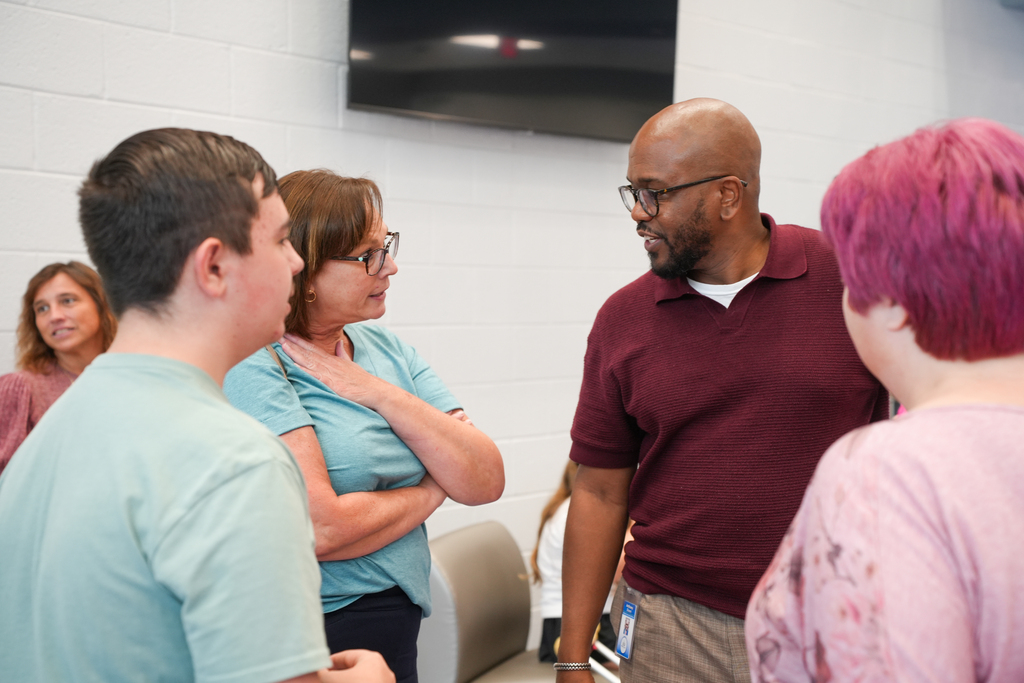 A group of people converse in a room with white walls. A man and woman in the center face each other.