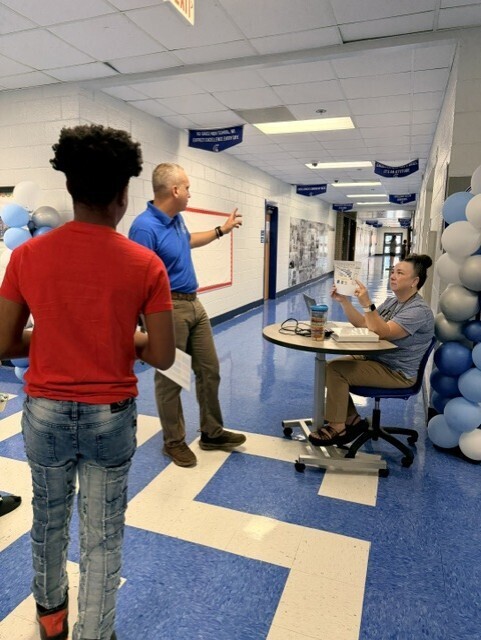 Two people in a hallway. One stands with a paper, the other seated at a desk. Both have blue and white balloons in the background.