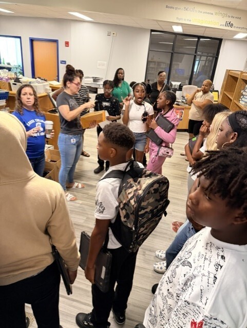 A group of children and adults in a classroom setting, some with backpacks, gathered around a woman.