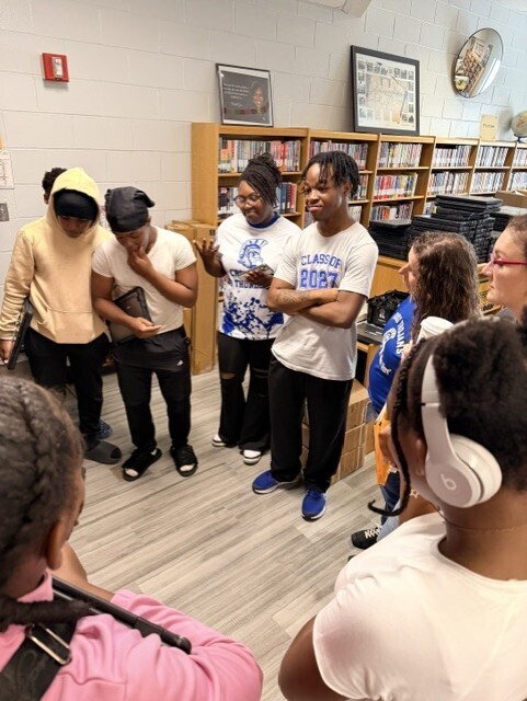 Group of students in a library, some wearing white shirts, some wearing headphones. One student holds a book.