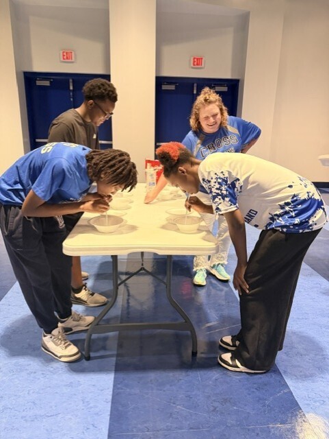 Four individuals lean over a table with bowls, likely participating in a food challenge. Two wear blue shirts, one in white.