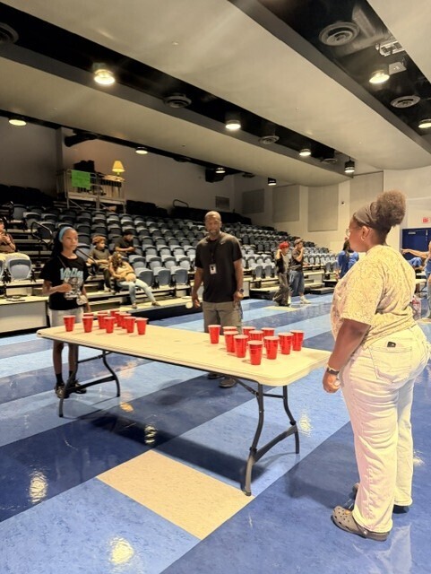 Two people standing at a long table with red cups, others around, an empty auditorium in the background.