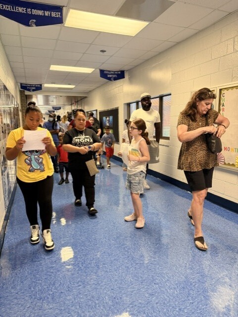 People, including adults and children, walk down a hallway. Some hold papers, and one adult wears a hat.