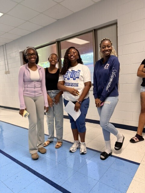 Five young women in a hallway, one holding paper, two others standing, blue floor, white walls.
