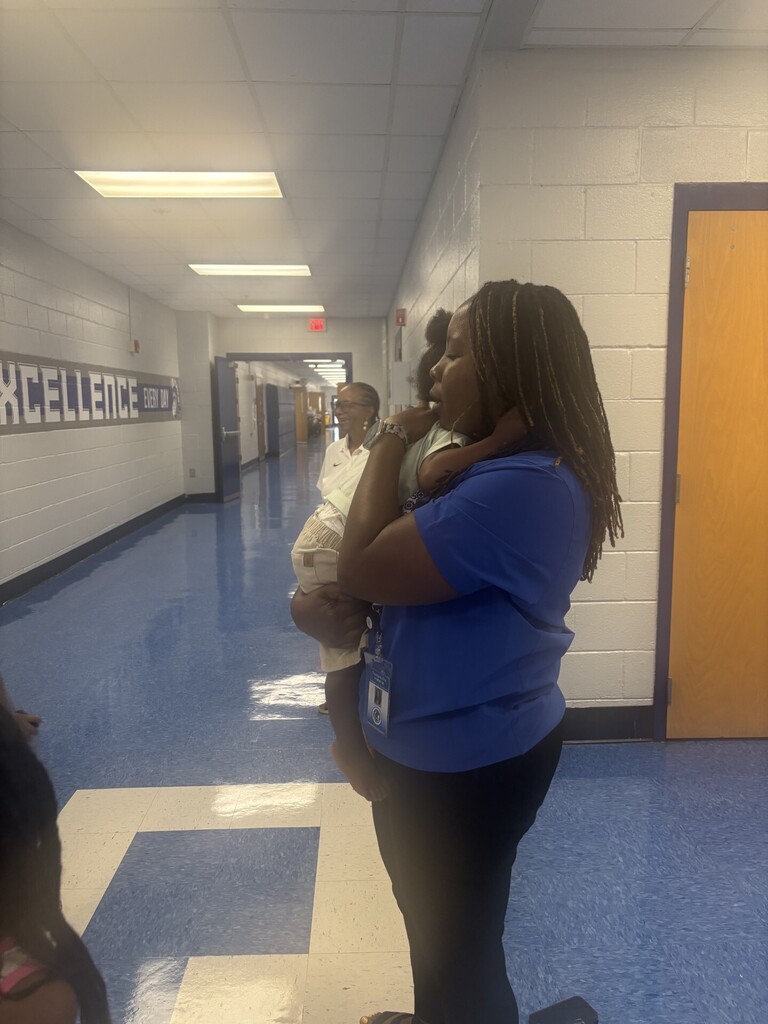 A woman holds a child in a school hallway. The walls are white, and the floor is blue.