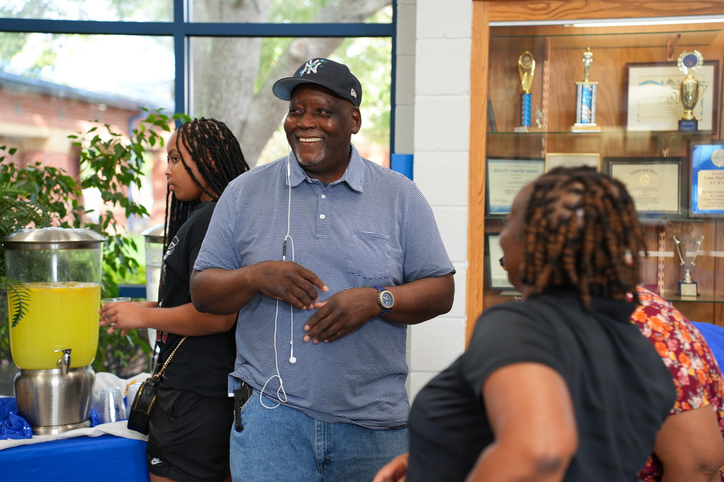 A man with a cap smiles, while two women listen. Behind them, a glass window and a display case with trophies.