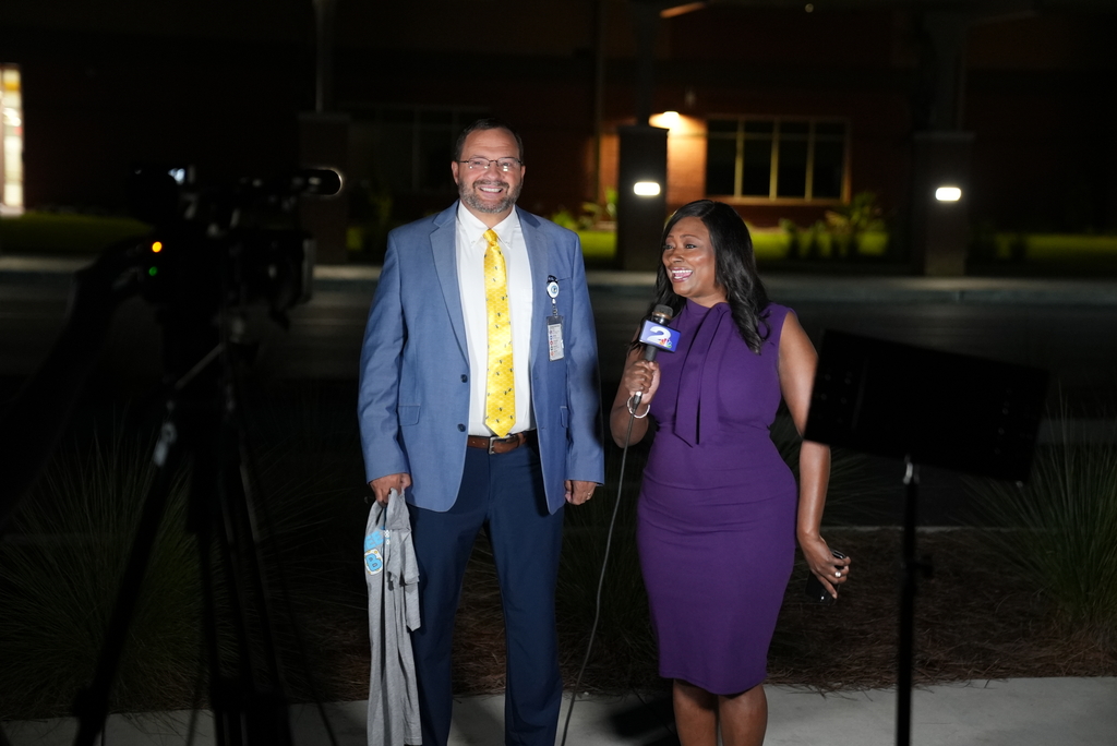 Man and woman stand outdoors at night, with a camera on a tripod behind them. The woman holds a microphone.