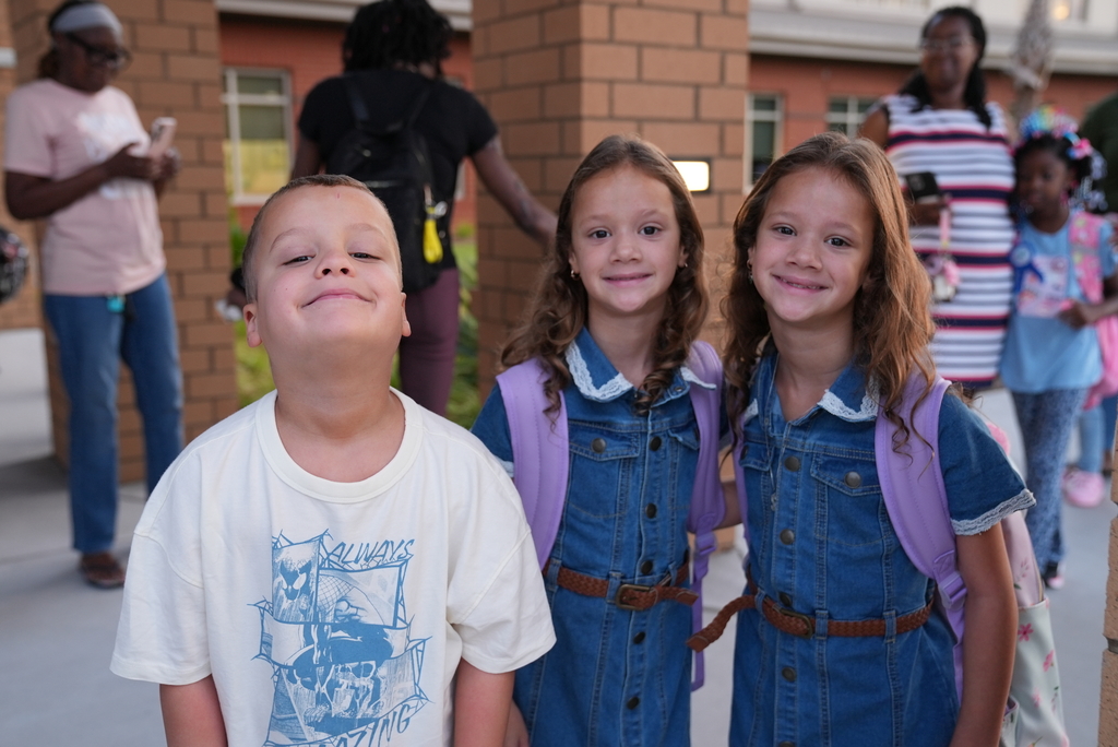 Three children, two girls and a boy, smile and stand together in front of a building.