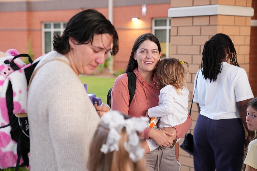 Several people standing outside a building. Two women, one with a baby, smile at a girl. A woman with a pink backpack stands nearby.