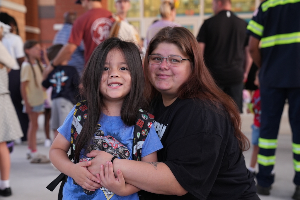 A woman with glasses holds a smiling child wearing a backpack in a hallway with people.