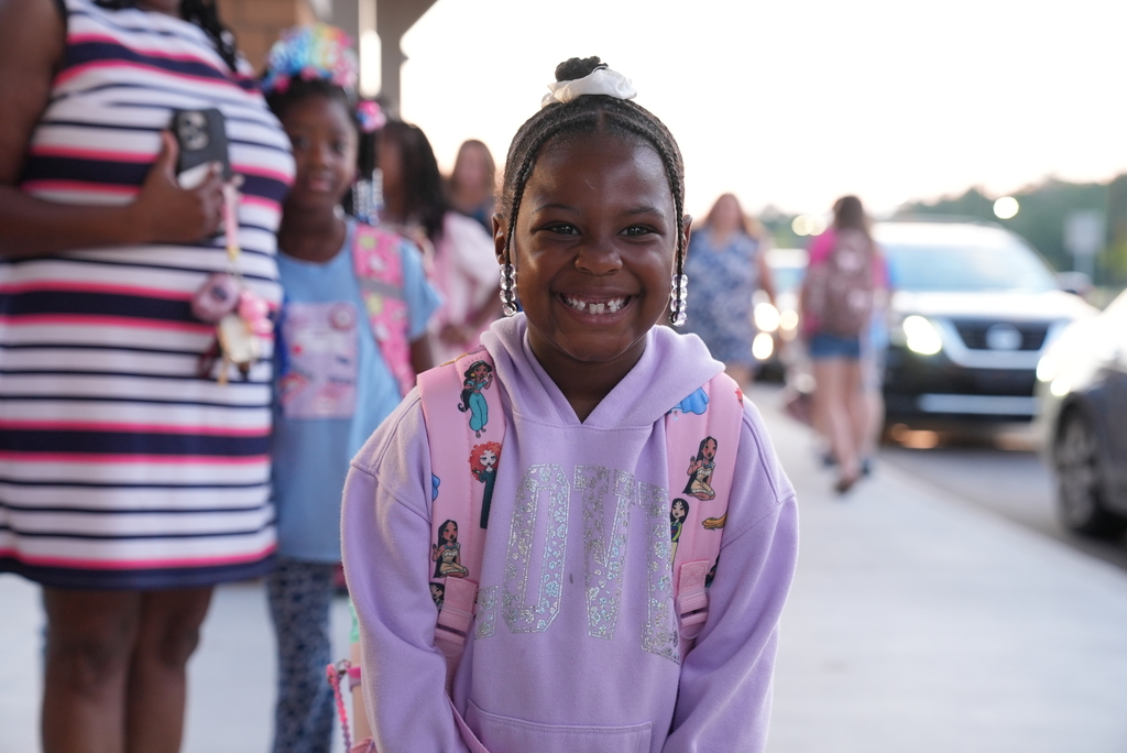 A smiling girl with a backpack stands on the sidewalk with other children, some wearing backpacks.