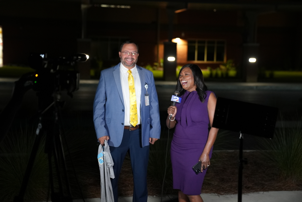 A man and a woman stand outside at night. The man holds a jacket. The woman holds a microphone and smiles.