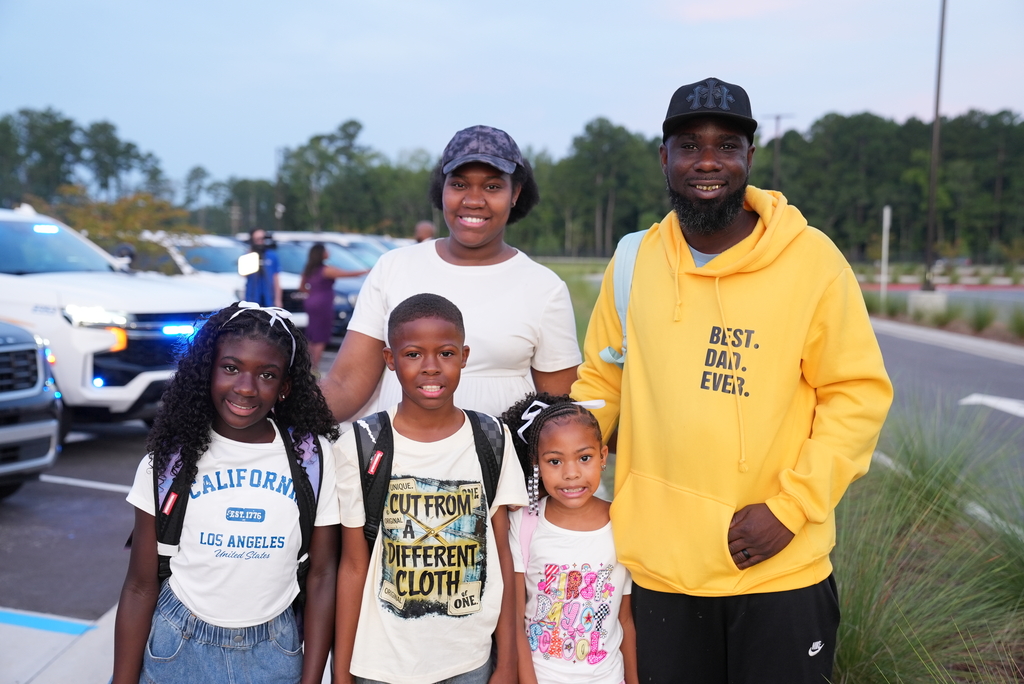 A family stands in a parking lot with parked cars and trees in the background.