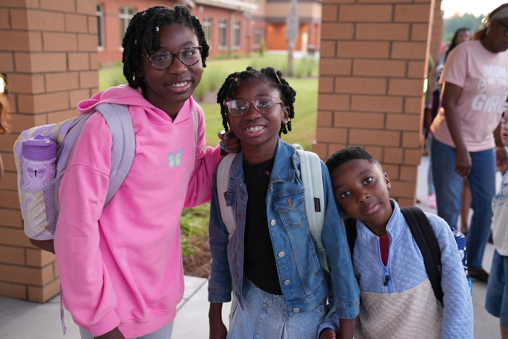 Three kids standing close together, two girls and a boy, smiling. Background includes a building with brick walls.