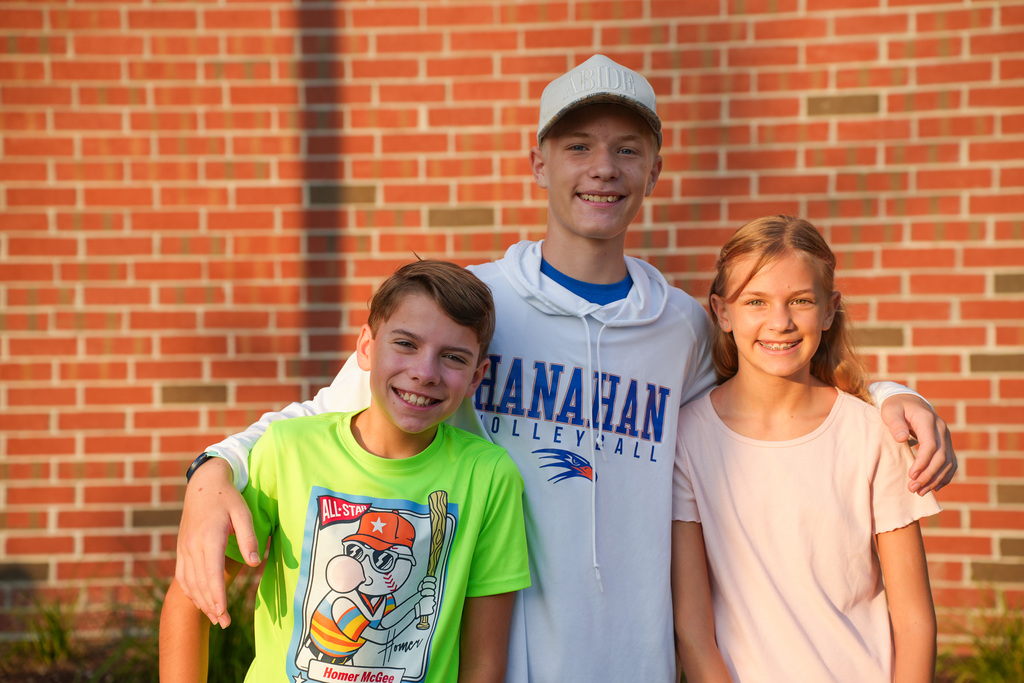 Three kids standing in front of a brick wall, smiling. The boy in the middle wears a green shirt.