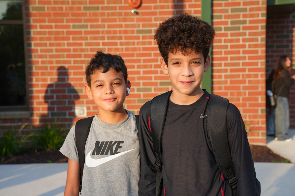 Two boys, one in a gray shirt with Nike, the other in a black shirt, stand smiling before a brick building.