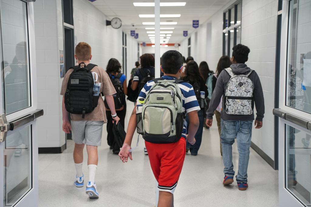 A group of students walk in a school hallway with white walls, glass doors, and backpacks.