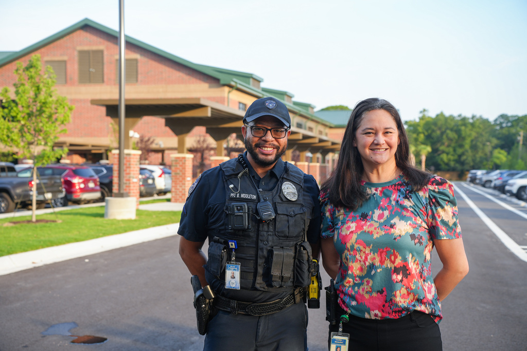 A police officer and a woman stand in a parking lot, smiling, with a building and parked cars behind them.