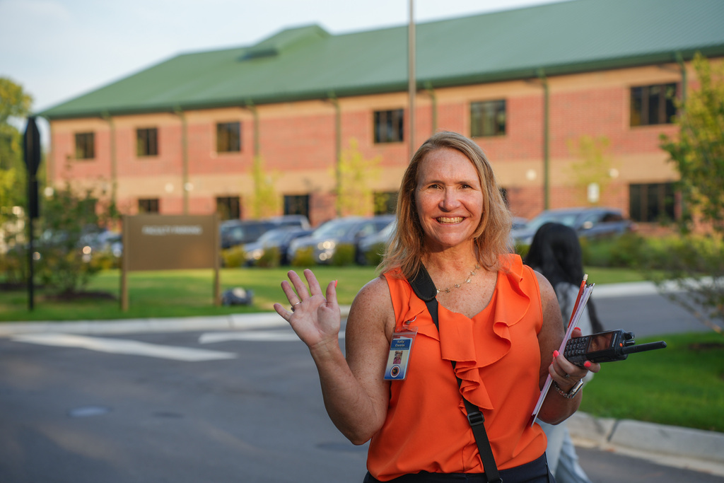 Woman in orange shirt and black pants waves while holding a folder and a remote control. Behind her is a building with a green roof.