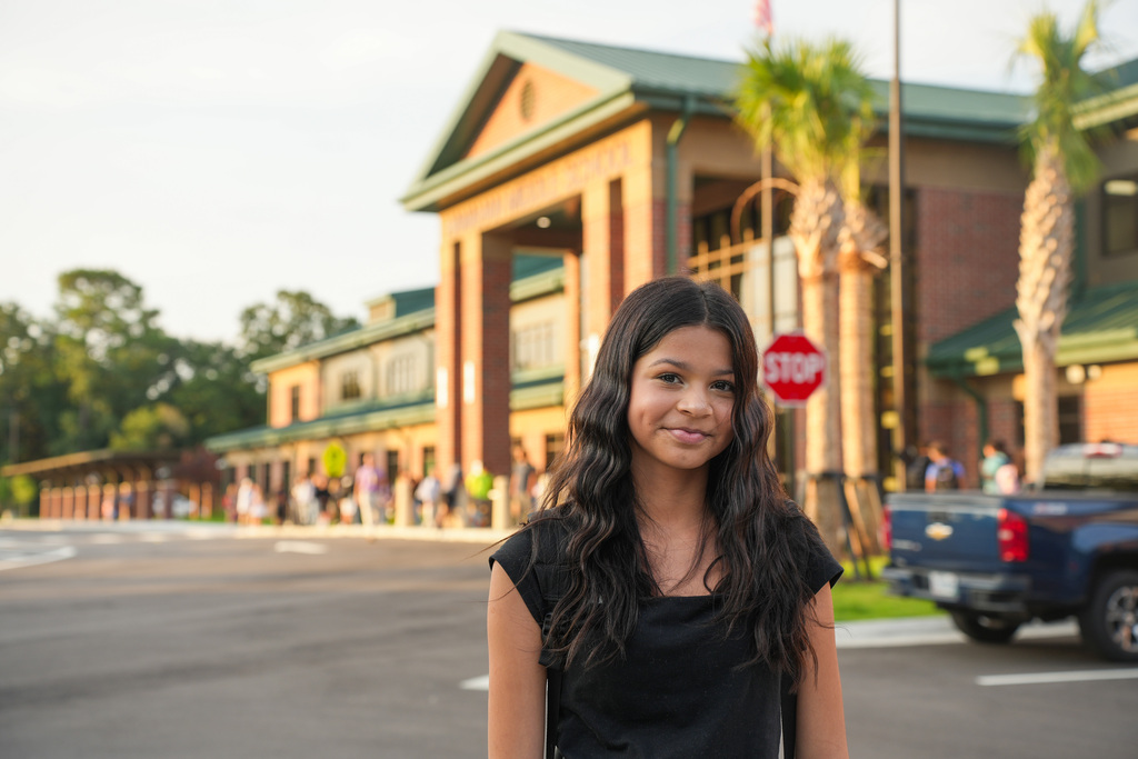 A girl with long dark hair stands in a parking lot. Behind her is a building with a green roof and palm trees.