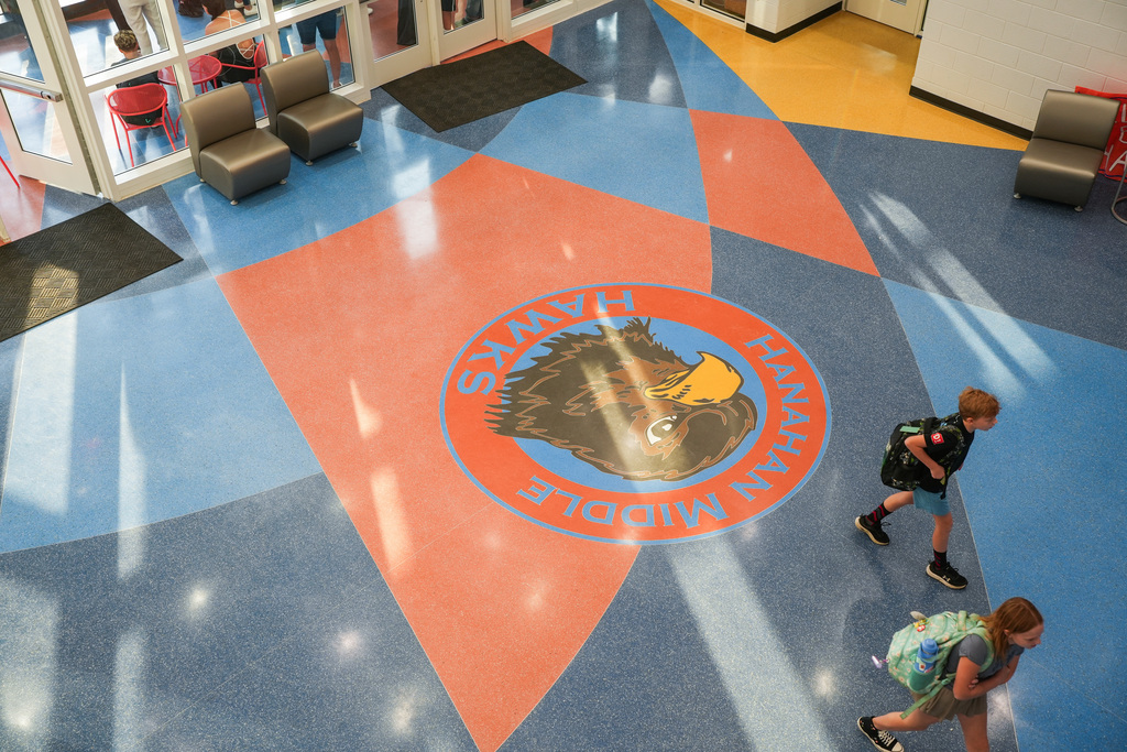 A school hallway features a large logo on the floor. Two individuals walk by, one carrying a backpack.