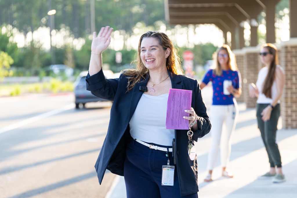 A woman waves while walking on a sidewalk with two other women; one holds a purple folder. A car drives by on the road.