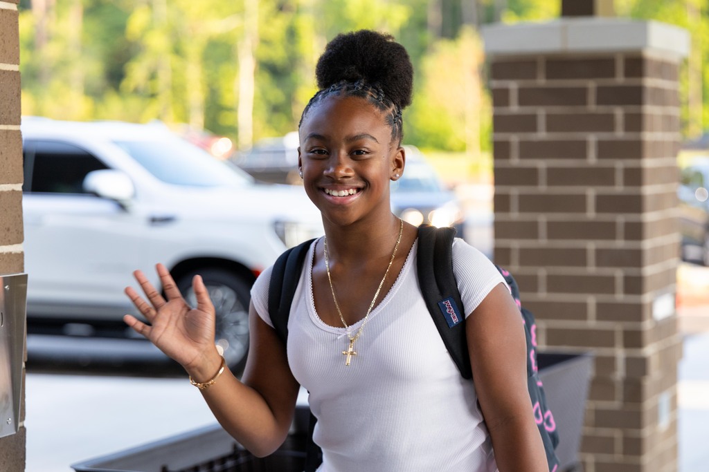 Person wearing a white t-shirt, a necklace, and a backpack standing in front of a brick wall.