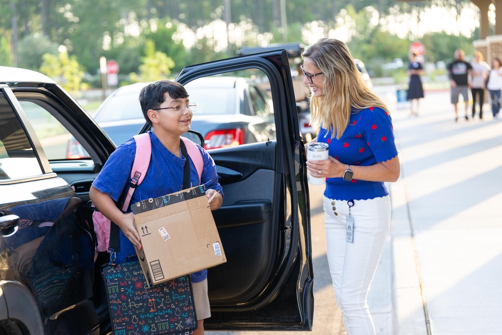 A person with glasses and a backpack helps another person with a cardboard box outside near a parked car.