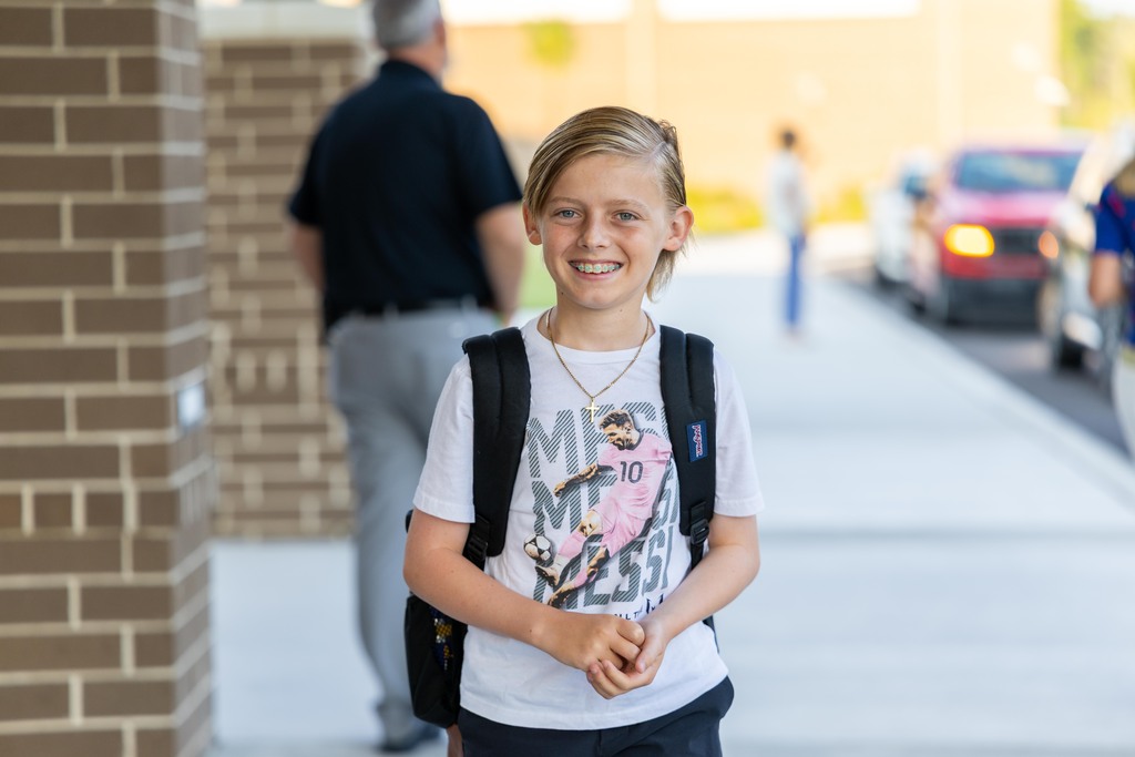 A child stands on a sidewalk, smiling and wearing a white T-shirt, a backpack, and a necklace.
