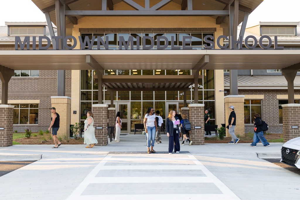 People walk near a building. The name reads "MIDDOW MIDDLE SCHOOL." A car is parked.