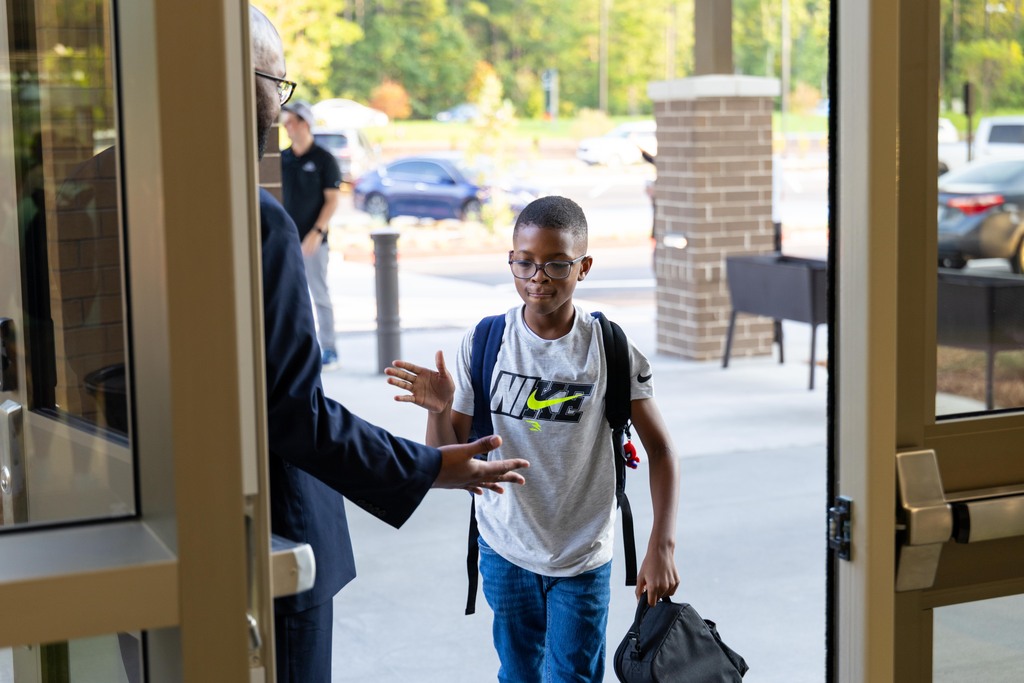 A young boy in a white t-shirt, eyeglasses, and a backpack walks out of a door. A man in a suit gestures as the boy exits.
