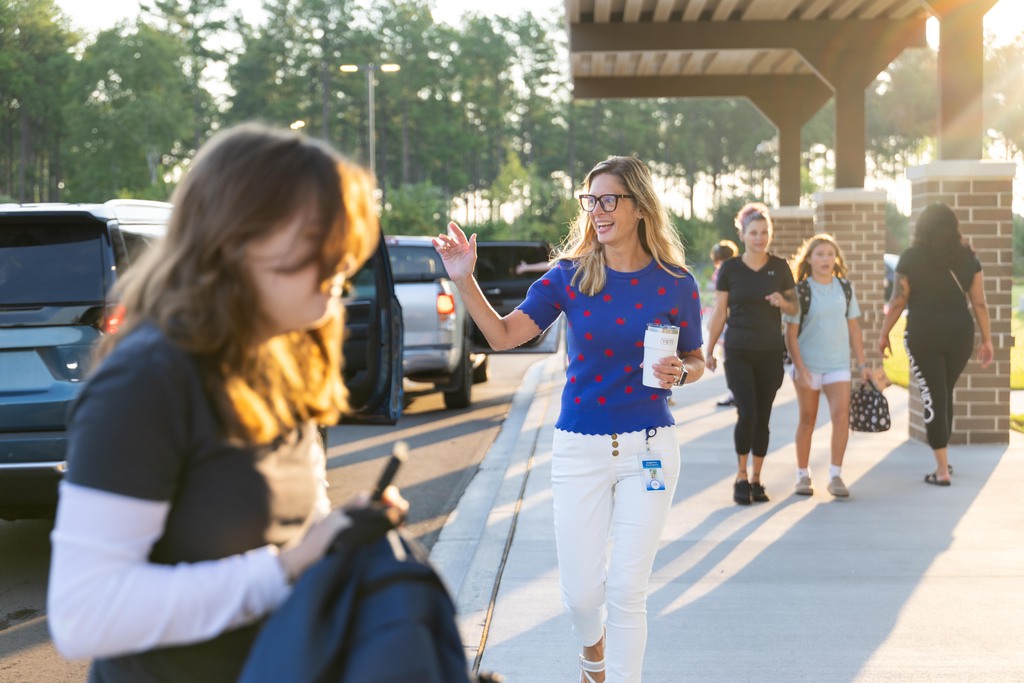 A woman speaks to another woman with a backpack. A woman in blue waves, and people walk by.