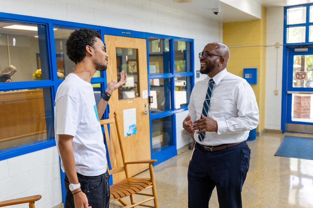 Two men stand in a room with blue trim, one gesturing. Behind them, a wooden chair, glass windows, and a door.