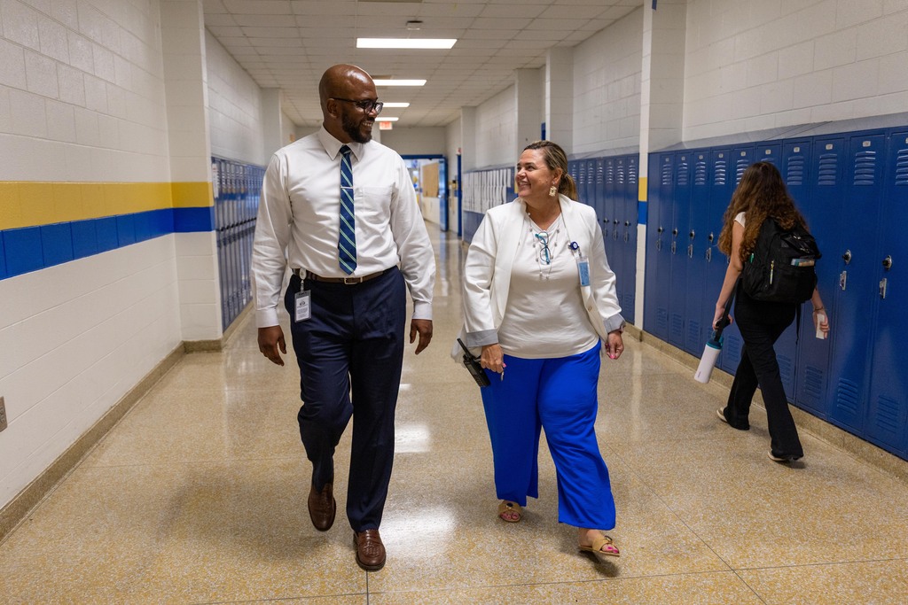 Two people in professional attire walk through a hallway. One wears a white shirt and blue pants, the other in white and blue. Another person walks behind them.