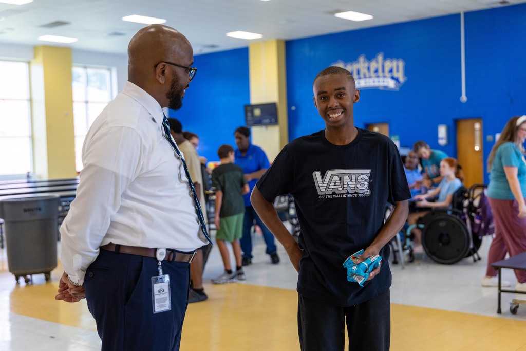 Two men stand in a school hallway with blue walls, one wearing a tie and the other a Vans shirt.