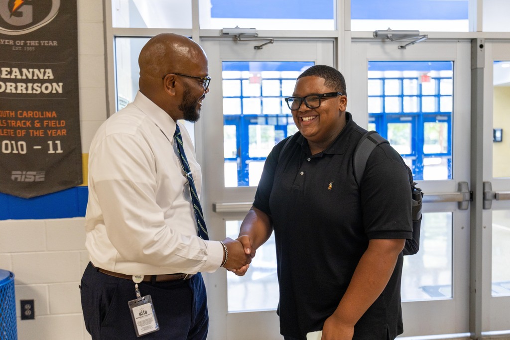 Two men, one in a white shirt and tie, the other in a black shirt with a backpack, shake hands.