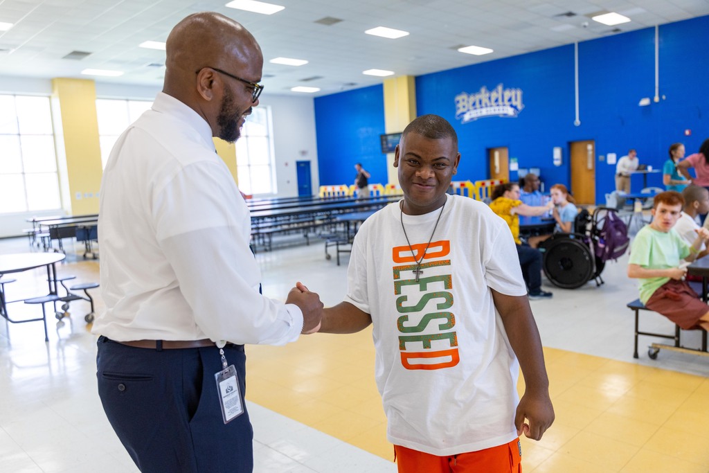 Two men shake hands in a cafeteria. One wears a white shirt, and the other wears orange pants. Behind them, people sit at tables and near doors.