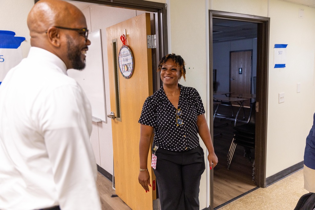 Two people converse near an open door; a man in a white shirt and a woman in a polka-dot shirt.