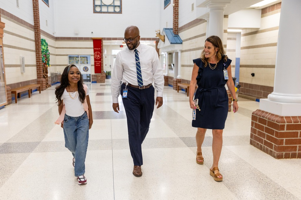 A man and two women walk down a school hallway. The man wears a white shirt and tie, and the women wear dark dresses.