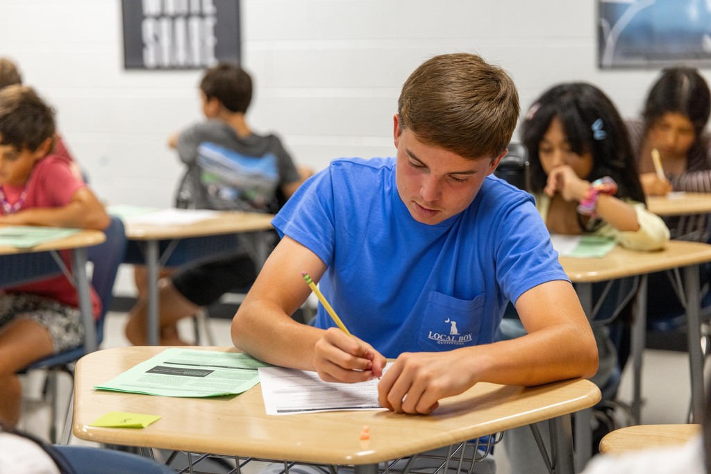 A student in a blue shirt is writing on paper at a desk in a classroom.