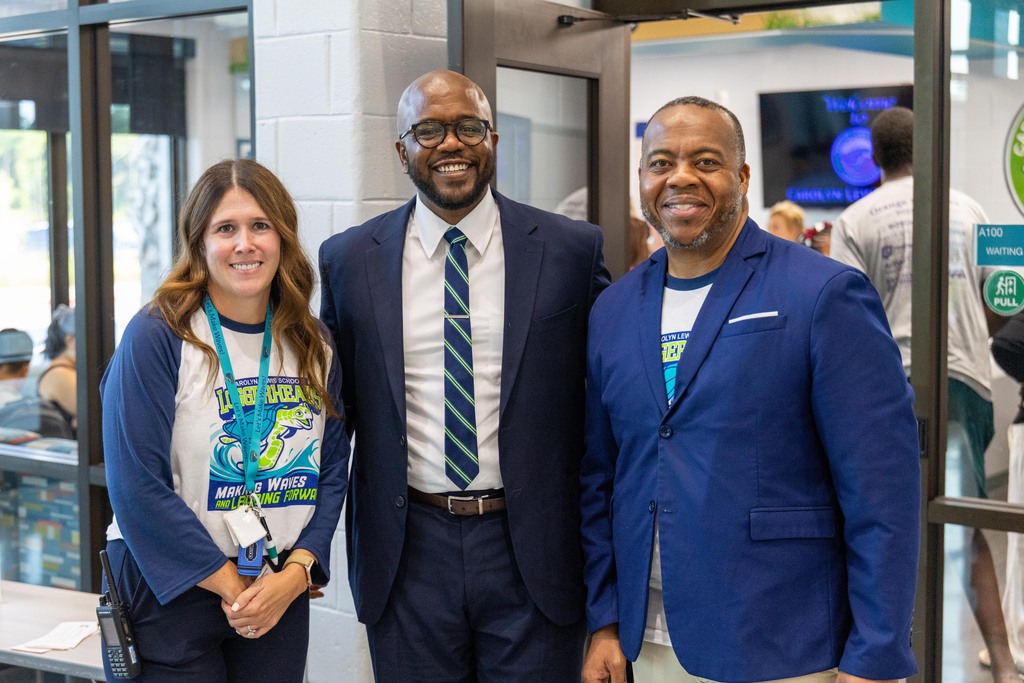 Three people stand together, smiling for a photo. One wears a white shirt with a blue lanyard, another in a blue suit and tie.