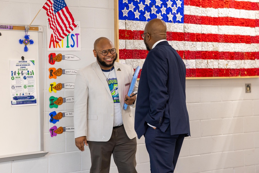 Two men stand close, one holds a folder. Behind them, a flag and whiteboard with colorful charts.