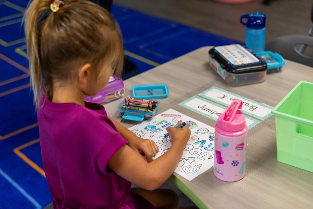 A young girl in a purple shirt sits at a table and colors a worksheet, with crayons, a bottle, and other supplies nearby.