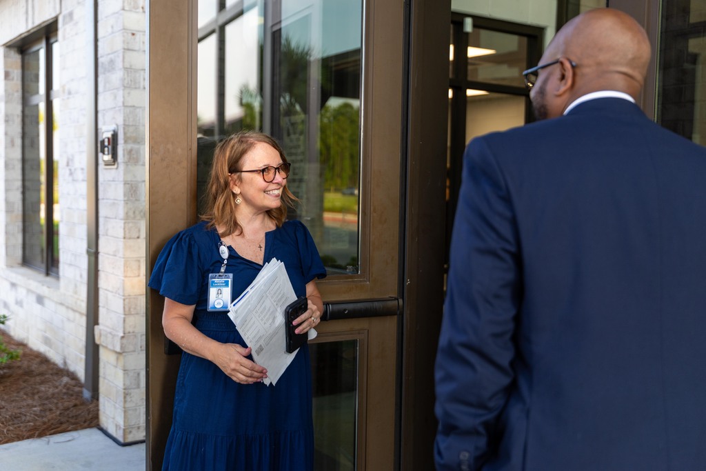 A woman in a blue dress holds papers in front of a building. A man in a blue suit and glasses stands beside her.